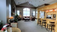 This inviting single-floor space is designed to foster comfort and community, combining architectural elegance with warm, functional furnishings. The centerpiece of the room is its soaring vaulted ceiling, accented by a ceiling fan and crown molding details that add a refined touch. A striking stone fireplace anchors one side of the space, providing a cozy focal point framed by lush indoor plants that bring a natural, homey feel. Abundant natural light pours in through multiple large windows and a prominent arched window at the far wall, creating a bright and airy atmosphere throughout. The main seating area features a generous sectional sofa and armchairs arranged around a central coffee table, offering ample room for socializing or unwinding. To one side, a built-in wooden bar with bar-height seating provides a casual gathering spot, complete with overhead cabinetry and pass-through counter space. Neutral carpeting runs throughout, softening the open floor plan and contributing to the space's overall warmth. The layout flows openly and efficiently, making the area well-suited for a variety of uses, from quiet relaxation to organized community events.