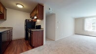 Located at 1604 Waunona Way in Madison, Wisconsin, this single-floor unit offers a practical and comfortable layout designed for everyday living. The kitchen serves as a functional centerpiece, featuring rich, dark-stained wood cabinetry with ample upper and lower storage, granite-patterned countertops, and black appliances including a refrigerator, gas range, and built-in microwave. Hardwood-style flooring runs throughout the kitchen, providing a warm contrast to the neutral-toned carpet that extends into the adjoining living and bedroom areas. The galley-style kitchen layout maximizes efficiency, with countertops and cabinetry lining both sides and opening directly into the living space.

The living area is open and well-proportioned, featuring light-colored walls and soft carpeting that create a calm, neutral ambiance. The bedroom is generously sized, bathed in natural light from a window with blinds, and equipped with a baseboard heating unit along the wall. The bathroom includes a full soaking tub with tiled surround in warm earth tones, a vanity with dark wood cabinetry and a light countertop, and tile flooring. Overall, the unit presents a clean, straightforward aesthetic throughout.