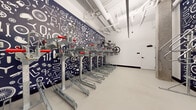 This single-level indoor bicycle storage room, located in Hayward, CA (94544), is a purpose-built space designed to accommodate a high volume of bicycles in an organized and efficient manner. The room's most striking visual feature is its bold, navy blue accent wall adorned with a white illustrated mural depicting bicycles, tools, gears, and cycling components — a thematic design choice that reinforces the room's dedicated function and adds a dynamic, urban character to the otherwise clean, utilitarian space.

The storage system consists of rows of heavy-duty galvanized steel two-tier bike racks, each equipped with red-handled pull-out mechanisms that allow bicycles to be stored on both the lower and elevated upper levels, maximizing the use of vertical space. The racks are arranged in parallel rows, providing clearly defined storage positions throughout the room. The polished concrete-style flooring is smooth and durable, well-suited for rolling bikes in and out. Exposed overhead pipes run along the white ceiling, contributing to the industrial aesthetic of the space.

A secured entry door provides controlled access, ensuring the safety of stored bicycles. Structural concrete columns are visible, reflecting the building's robust construction. The room's clean white walls and bright lighting create a well-lit, functional environment that is both practical and visually engaging for daily users.