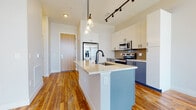This well-appointed single-floor unit offers a thoughtfully designed open-concept layout that seamlessly connects the kitchen and living areas. The kitchen serves as the centerpiece, featuring a large island with a white quartz countertop, an integrated sink, and a matte black faucet. Two-tone cabinetry — crisp white upper cabinets paired with slate blue lower cabinets — adds a contemporary aesthetic, while a neutral tile backsplash provides a refined backdrop for the stainless steel appliances, including a French door refrigerator, electric range, and over-the-range microwave. Pendant lighting with glass shades and a black track light rail illuminate the kitchen, complementing the warm, richly grained hardwood flooring that extends throughout the main living area. The bathroom is finished with large-format grey tile flooring, white subway-tiled tub surround, and coordinating matte black hardware, creating a clean and modern feel. The bedroom is carpeted for comfort and benefits from floor-to-ceiling windows that flood the space with natural light and offer sweeping views of the surrounding cityscape. Throughout the unit, a neutral color palette and modern fixtures work together to create a cohesive, contemporary living environment.
