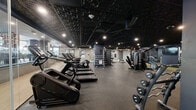 Located at 51 South Broadway in White Plains, NY, this fitness facility offers a spacious, single-floor layout thoughtfully designed to accommodate a full range of workout needs. The interior features a striking dark aesthetic, with a textured black ceiling, recessed spotlighting, and deep navy accent walls that create a focused, high-energy atmosphere throughout the space.

The main workout floor is divided into distinct functional zones. A robust cardio area lines one side of the facility, equipped with StairMaster climbers, treadmills, and elliptical machines. The strength training section features an extensive dumbbell rack with a wide range of free weights, adjustable benches, and multiple cable and selectorized machines, including a cable motion system and various resistance stations. A dedicated free weights area with squat racks and barbell equipment supports powerlifting and compound movements.

A separate room with glass-panel walls and a lighter color palette provides a more intimate space for stretching, functional training, or individual workouts, and includes mirror walls that enhance the sense of openness. Rubber flooring throughout ensures safety and durability, while full-length mirrors along key walls allow for proper form checks. Motivational black-and-white photography adds character to the space. A water fountain is conveniently accessible near the secondary room.