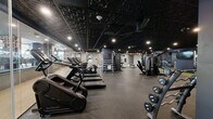 Located at 51 South Broadway in White Plains, NY, this fitness facility offers a spacious, single-floor layout thoughtfully designed to accommodate a full range of workout needs. The interior features a striking dark aesthetic, with a textured black ceiling, recessed spotlighting, and deep navy accent walls that create a focused, high-energy atmosphere throughout the space.

The main workout floor is divided into distinct functional zones. A robust cardio area lines one side of the facility, equipped with StairMaster climbers, treadmills, and elliptical machines. The strength training section features an extensive dumbbell rack with a wide range of free weights, adjustable benches, and multiple cable and selectorized machines, including a cable motion system and various resistance stations. A dedicated free weights area with squat racks and barbell equipment supports powerlifting and compound movements.

A separate room with glass-panel walls and a lighter color palette provides a more intimate space for stretching, functional training, or individual workouts, and includes mirror walls that enhance the sense of openness. Rubber flooring throughout ensures safety and durability, while full-length mirrors along key walls allow for proper form checks. Motivational black-and-white photography adds character to the space. A water fountain is conveniently accessible near the secondary room.