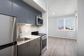 This thoughtfully designed single-floor unit offers a clean, modern aesthetic throughout, with light hardwood-style flooring and neutral wall tones creating a bright, cohesive atmosphere. The galley-style kitchen is efficiently laid out and well-appointed, featuring sleek grey cabinetry, stainless steel appliances including a full-size refrigerator and range, a stainless undermount sink with a modern pull-down faucet, and a classic subway tile backsplash. Recessed lighting illuminates the workspace effectively. The kitchen opens toward a windowed living area that benefits from natural light, contributing to an airy, open feel. The bathroom is clean and functional, finished in soft neutral tones with a white vanity, sink, toilet, and a shower/tub combination with a mirror above. A standout convenience feature is the dedicated in-unit laundry closet, which houses a stacked washer and dryer unit accessible via a sliding door. An additional room provides flexible living space, with a sliding closet door and connections to adjacent areas of the unit. The overall design prioritizes smart use of space, modern finishes, and practical amenities, making it a well-equipped and move-in-ready residence.