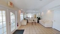 Located at 5461 Collins Mill Creek Drive in Milton, Florida, this single-story space offers a welcoming and well-appointed interior designed for comfort and functionality. The main living area is a standout feature, boasting vaulted ceilings with a ceiling fan that enhance the sense of openness and airflow throughout the room. Large arched windows along the front wall flood the space with generous natural light, creating a bright and airy atmosphere. Light-toned ceramic tile flooring extends across the living area, offering a clean and cohesive look that complements the neutral white walls and trim. A full-wall mirror is positioned at one end of the living room, visually expanding the space and reflecting the natural light from the windows. Recessed lighting supplements the natural illumination, ensuring the interior remains well-lit throughout the day. The textured popcorn ceiling adds a classic architectural detail consistent with the home's overall style. A secondary room is visible through an arched doorway, suggesting a versatile floor plan that accommodates multiple uses. The overall ambiance of this space is light, open, and inviting, with a clean, neutral palette that lends itself well to a variety of interior design styles.