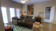 This single-floor residence offers a thoughtfully designed living space that balances comfort and style. The living area serves as the heart of the home, featuring warm neutral walls that create a welcoming, relaxed atmosphere. A ceiling fan with a light fixture anchors the room from above, ensuring both comfort and functionality. Large French doors or wide windows along one wall flood the space with abundant natural light, enhancing the open and airy feel of the interior. The room is furnished with a spacious gray sofa accented with green and neutral throw pillows, complemented by a matching armchair and a pair of upholstered ottomans in a classic patterned fabric. A dark wood coffee table with a lower storage shelf serves as a central gathering point, while coordinating end tables with warm-toned lamps add a balanced, symmetrical touch to the layout. A bold nature-inspired artwork depicting a tree-lined path adds visual depth and a sense of the outdoors to the wall above the sofa. The vivid green geometric-patterned area rug grounds the seating arrangement and introduces a lively pop of color. Together, these elements create a cohesive, inviting living environment that is both practical and visually appealing.