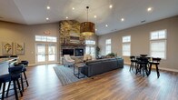 The Ashton Park clubhouse is a thoughtfully designed single-floor community space that combines modern aesthetics with comfortable, functional living. The centerpiece of the main lounge is a dramatic floor-to-ceiling stacked stone fireplace, flanked by large windows and glass double doors that flood the area with natural light and provide views of the outdoor surroundings. Soaring vaulted ceilings amplify the sense of openness, while oversized drum pendant lights add a warm, contemporary accent. Rich hardwood flooring flows seamlessly throughout the entire space, anchoring the neutral warm-gray palette on the walls.

The lounge area offers generous seating with a large sectional sofa and accent chairs arranged around a patterned area rug. Adjacent to the lounge, a dining and bar-height seating area provides additional gathering space, decorated with botanical artwork. The fully equipped kitchen features dark shaker-style cabinetry, granite countertops, stainless steel appliances, and a curved island, making it ideal for hosting events.

Toward the rear of the space, a quieter sitting area leads to dedicated private office rooms with glass-panel walls, offering workstations for focused tasks. The overall ambiance is polished, welcoming, and well-suited for both social gatherings and productive work.