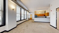 Located in Cleveland's vibrant West Ninth Street corridor, this two-bedroom, two-bathroom residence blends industrial character with comfortable living. The open-concept living area immediately impresses with a wall of large, black-framed windows that flood the space with natural light and offer street-level views of the surrounding neighborhood. Exposed cylindrical ductwork runs along the ceiling, lending an authentic loft aesthetic throughout the main living area.

The kitchen is positioned adjacent to the living space, separated by a raised breakfast bar. It features warm honey-toned cabinetry, dark granite countertops, tile flooring, and a full suite of black appliances including a refrigerator, range, and over-the-range microwave.

The primary bedroom is generously proportioned, offering ample space and large windows with the same bold black framing found throughout the unit. The second bedroom is more intimate, with carpeted flooring and a window that brings in natural light. Both bathrooms are consistently appointed with dark granite vanity countertops, warm wood cabinetry, tiled flooring, and full shower-tub combinations. Soft carpeting runs throughout the living and bedroom areas, adding warmth to the industrial-inspired design.