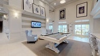 This expansive two-floor clubhouse is designed in a refined coastal aesthetic, blending bright, airy interiors with thoughtfully curated furnishings throughout. The main floor opens into a grand common area with soaring double-height ceilings, exposed structural beams, large-format white tile flooring, and a mezzanine level accessible by a central staircase with glass-panel railings, flooding the space with an impressive sense of openness. A dedicated billiards room anchors one wing, featuring a statement white pool table, soft blue wingback chairs, shiplap accent walls with horizontal stripe detailing, and oversized coastal-themed artwork displayed at height. An adjoining dining area offers a large rustic wood trestle table surrounded by ticking-stripe upholstered chairs, complemented by a wall-mounted TV and a wet bar with beverage refrigeration. A formal conference-style dining room presents bold cobalt blue seating beneath elegant lantern pendant lights with a mounted display screen. The catering kitchen is fully outfitted with stainless steel double wall ovens, built-in microwave, panel-ready refrigeration, ample grey cabinetry, and white quartz countertops. A media lounge with tiered seating and a ceiling-mounted projector provides a private screening experience. The lower floor continues the coastal theme with dark hardwood-style flooring, built-in cabinetry, gallery wall displays, and flexible lounge seating.