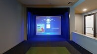 Located in Richmond, VA, this single-floor indoor golf simulator facility offers a sleek, purpose-built environment designed for both performance and comfort. The space is divided into two primary areas that work seamlessly together to deliver a complete golf simulation experience.

The simulator bay is the centerpiece of the facility, housing a Full Swing golf simulator complete with a large projection screen, a synthetic turf hitting mat, and striking blue ambient lighting that creates an immersive, focused atmosphere. Dark carpeting and black-trimmed walls within the bay enhance contrast and reduce visual distraction during play. A wall-mounted control monitor adjacent to the bay provides easy access to simulator settings and game selection.

The adjacent lounge and reception area features light-toned tile flooring, soft gray walls, and recessed ceiling lighting that establishes a clean and welcoming environment. Contemporary seating, including woven accent chairs and small side tables, offers a comfortable space for waiting or observing. A bold abstract canvas print adds a touch of artistic character to the walls. A workstation counter is also present, supporting a professional setup suitable for client check-ins or consultations. A window dressed with tailored drapes and blinds allows natural light to filter into the lounge area. Golf equipment storage is visible near the window, reinforcing the facility's dedicated, sport-focused purpose.