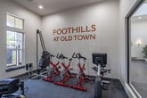 Located at 28845 Pujol St in Temecula, CA, this single-floor fitness center at Foothills at Old Town offers a comprehensive workout facility designed to support a wide range of exercise routines. The main gym area is generously proportioned, featuring soaring ceilings and an abundance of natural light streaming through large, multi-pane windows that offer views of the surrounding landscaped grounds. Multiple ceiling fans are mounted overhead to maintain comfortable airflow throughout the space. The main floor is outfitted with a variety of cardio equipment, including treadmills, elliptical trainers, and rowing machines, all positioned to take advantage of the bright, window-lined walls. Strength training equipment, including a cable machine and weight bench, occupies a dedicated section of the gym. A separate enclosed room serves as a cycling and specialty equipment studio, housing spin bikes and a curved treadmill, with branded wall signage and a wall-mounted television for an immersive workout experience. The facility also includes a well-appointed hallway with gender-designated restrooms and a dual-basin water fountain with a bottle-filling station. Durable carpet tile flooring throughout provides traction and comfort, and multiple wall-mounted televisions ensure entertainment options are always within view.