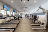 Located at 3089 James Ct in Lake Charles, Louisiana, this fitness center offers a thoughtfully designed workout space accommodating a full range of exercise needs. The single-floor layout is efficiently organized, providing generous room to move between stations without feeling cramped.

The cardio section features a row of Life Fitness treadmills and elliptical machines lined along one side of the facility, each equipped with individual screens for entertainment during workouts. Multiple wall-mounted flat-screen TVs are positioned throughout the space for additional viewing convenience.

The strength training area is equally well-appointed, with a selection of Life Fitness selectorized machines, free weight benches, a dumbbell rack, and functional accessories such as medicine balls and stability balls. A large wall mirror along the back enhances the sense of space while allowing users to monitor their form.

The interior design is clean and modern, with white walls accented by a distinctive horizontal slate-blue stripe, recessed lighting, and large ceiling fans that promote air circulation throughout. Large windows along the perimeter bring in natural light and provide exterior views. Durable large-format tile flooring grounds the space with a practical, polished finish. The overall ambiance is bright, organized, and purpose-built for an effective fitness experience.