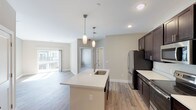 This single-floor residence features a thoughtfully designed open-concept layout with a neutral color palette and wood-look flooring throughout the main living areas. The kitchen serves as the centerpiece, equipped with rich dark espresso cabinetry, light quartz countertops, and a full suite of stainless steel appliances including a refrigerator, range, and over-the-range microwave. A central island with an undermount sink and pendant lighting above provides additional workspace and a natural gathering point. Recessed lighting complements the pendants, keeping the space bright and functional. The kitchen opens directly into a spacious living area, which benefits from natural light through a sliding glass door leading to an outdoor balcony. The bedrooms feature plush wall-to-wall carpeting, ceiling fans, and generously sized windows with blinds that allow ample natural light while maintaining privacy. The bathroom includes a full tub-shower combination with a curved curtain rod, wood-look tile flooring, dark vanity cabinetry, and clean, understated finishes consistent with the rest of the unit. Overall, the space presents a cohesive, contemporary aesthetic with practical finishes suited for everyday living.