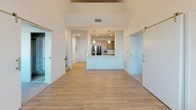 This two-story residence showcases a thoughtfully designed open-concept layout with a bright, neutral palette and contemporary finishes. The main living area is defined by dramatically high ceilings and light-toned hardwood-style flooring, creating an airy, expansive atmosphere. Sliding barn doors on hardware rails add a modern architectural accent, while large windows invite natural light and outdoor views.

The kitchen is centrally positioned as an island-style hub, featuring granite-look countertops, warm wood cabinetry, stainless steel appliances including a gas range and refrigerator, and pendant lighting overhead. Tile flooring anchors the kitchen zone distinctly from the surrounding living space.

The upper level includes a loft-style area with hardwood-style flooring, angled walls, and ceiling vents, offering flexible use as an office or additional living space. Bedrooms feature soft carpet flooring and windows with outdoor views.

Bathrooms are finished in a clean, modern style with light wood vanity cabinetry, white countertops, large-format tile flooring, and well-lit vanity mirrors. Chrome fixtures and open shelving complete the refined, functional aesthetic throughout.
