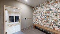 Located in Miami, FL, this wellness sauna suite is a compact, thoughtfully designed space composed of two distinct areas: an antechamber and a private infrared sauna room. The antechamber serves as a transitional space where visitors can prepare before and unwind after a session. A striking floor-to-ceiling accent wall clad in vibrant botanical tiles — featuring tropical birds, leaves, and flowers in earthy greens, oranges, and whites — anchors the room with a bold visual focal point. A solid wood bench sits against this decorative wall, offering a place to rest. A row of coat hooks lines the neutral gray wall opposite, and a dark-framed glass door provides entry to the suite. Light gray floor tiles carry throughout, maintaining a clean, cohesive aesthetic. A dark-finished shelving unit near the entry provides organized storage for personal belongings. The infrared sauna room itself is fully lined in light natural pine wood — walls, ceiling, and tiered benches — creating a warm, enveloping atmosphere. Multiple black infrared heating panels are integrated into the walls, ensuring even heat distribution throughout the cabin. Recessed ceiling lighting illuminates both spaces with a soft, controlled glow, contributing to the calm, spa-like ambiance of the overall suite.