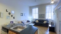 This single-floor space offers an open-plan layout that integrates the kitchen and living area into one cohesive environment. The kitchen is outfitted with a dark granite-style countertop and a stainless steel sink, providing a functional workspace that flows directly into the main living area. The living room is comfortably furnished with two sofas — a light-toned couch accented with navy blue pillows and a larger dark sectional with yellow accent cushions — arranged to encourage conversation and relaxation. A sleek marble-top coffee table anchors the seating arrangement. The room benefits from generous natural light streaming through multiple windows dressed with sheer curtains, creating a bright and airy atmosphere. The warm hardwood flooring throughout adds character and a sense of continuity between spaces. Tall ceilings enhance the feeling of openness, while the neutral wall tones provide a clean backdrop for the decorative touches, including wall art that adds personality to the space. The overall ambiance is casual yet inviting, making it a comfortable and practical everyday living environment.