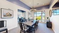 This well-appointed single-floor community space is divided into two interconnected rooms, seamlessly connected by elegant sliding barn doors that allow for flexible use and an open flow between areas. The design is anchored by a striking color scheme of deep periwinkle-blue upper walls contrasted against crisp white wainscoting and trim, creating a sophisticated and cohesive atmosphere throughout.

The primary room serves as a versatile dining and gathering area, highlighted by soaring ceilings that amplify the sense of space. A statement chandelier with exposed Edison-style bulbs hangs above a large rectangular dining table surrounded by rattan-accented chairs with striped cushions. Floor-to-ceiling windows with bold cross-mullion detailing flood the room with natural light and offer views of lush outdoor greenery, while patterned carpeting in soft blue tones grounds the space.

The second room functions as a business center, featuring a long built-in desk with multiple workstations, each equipped with blue ergonomic office chairs. A row of framed abstract artwork adorns the wall above the workstations, flanked by table lamps. A wall-mounted television and an additional table with seating provide further functionality. Together, these two rooms offer a refined and practical environment ideal for both social gatherings and productive work.