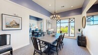 This well-appointed single-floor community space is divided into two interconnected rooms, seamlessly connected by elegant sliding barn doors that allow for flexible use and an open flow between areas. The design is anchored by a striking color scheme of deep periwinkle-blue upper walls contrasted against crisp white wainscoting and trim, creating a sophisticated and cohesive atmosphere throughout.

The primary room serves as a versatile dining and gathering area, highlighted by soaring ceilings that amplify the sense of space. A statement chandelier with exposed Edison-style bulbs hangs above a large rectangular dining table surrounded by rattan-accented chairs with striped cushions. Floor-to-ceiling windows with bold cross-mullion detailing flood the room with natural light and offer views of lush outdoor greenery, while patterned carpeting in soft blue tones grounds the space.

The second room functions as a business center, featuring a long built-in desk with multiple workstations, each equipped with blue ergonomic office chairs. A row of framed abstract artwork adorns the wall above the workstations, flanked by table lamps. A wall-mounted television and an additional table with seating provide further functionality. Together, these two rooms offer a refined and practical environment ideal for both social gatherings and productive work.