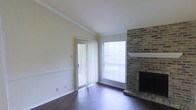 This single-story space presents a welcoming living area defined by a combination of classic architectural details and functional design. The room is anchored by a striking floor-to-ceiling brick fireplace, which serves as a prominent focal point and adds a warm, traditional character to the space. A white-painted mantel shelf accents the brick surround, providing a clean contrast against the natural masonry.

Dark hardwood floors run throughout the room, offering a rich, grounding aesthetic that complements the neutral, light-toned walls. Crown molding trims the ceiling, adding a refined architectural detail, while a chair rail along the lower portion of the walls contributes additional classic styling.

Natural light fills the space through two sources: a sliding glass door fitted with vertical blinds, which also provides access to an outdoor area, and an adjacent window with horizontal blinds. Together, these openings create a bright and airy atmosphere. The overall ambiance of the space is clean, neutral, and move-in ready, balancing traditional design elements with a simple, uncluttered layout suitable for a variety of furnishing styles.