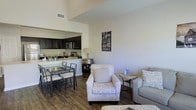 Located at 31611 Plaza De Toros Dr. in Spanish Fort, Alabama, this single-floor home offers an open-concept layout that seamlessly connects the main living areas. The kitchen is outfitted with dark cabinetry, stainless steel appliances, and a light-colored countertop that contrasts elegantly with the rich, dark wood-toned flooring found throughout. A pass-through design between the kitchen and living area enhances the sense of openness and flow. The dining area accommodates a glass-top table paired with classic metal chairs, positioned conveniently adjacent to the kitchen for easy entertaining. The living space features neutral-toned upholstered seating, including patterned accent chairs, creating a warm and inviting atmosphere. Notably high ceilings add to the sense of spaciousness, allowing natural light to fill the interior. Decorative touches, including a rustic barrel-style side table and framed wall art, contribute character and a relaxed, homey ambiance. The cohesive design throughout the space balances functionality with comfort, making this Spanish Fort home a well-appointed and welcoming single-level residence.