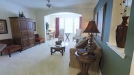 This single-floor residence showcases a refined traditional aesthetic with a seamless blend of comfort and classic elegance. The main living area is generously proportioned, featuring plush wall-to-wall carpeting and a ceiling fan overhead. Rich, dark wood furnishings — including a carved glass-topped coffee table and a round accent side table — anchor the space and contribute to its warm, sophisticated character. A large sofa with decorative pillows and a statement table lamp with an ornate base add layers of texture and detail. A striking arched passageway connects the main living area to a bright sitting or dining nook, creating an open and airy sense of flow throughout. This secondary space is bathed in natural light from a wide bay window dressed with bold red curtains, offering a welcoming spot for seating or casual dining. The accent wall in a soft teal tone provides a tasteful contrast to the neutral cream palette that predominates throughout. Framed artwork lines the walls, and thoughtful accessory placement throughout adds a curated, homey feel. The overall ambiance is warm, inviting, and elegantly appointed, ideal for both relaxed everyday living and comfortable entertaining.