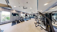 Located at 7560 Norman Rockwell Lane in Las Vegas, NV, this fitness center offers a comprehensive exercise environment designed to support a full range of workout routines. The space is thoughtfully laid out across a single floor, providing ample room for both cardio and strength training activities without feeling crowded.

The interior design features a distinctive geometric diamond-patterned accent wall in a navy and grey color palette, lending the space a modern, polished aesthetic. Light-toned flooring contrasts cleanly against the bold wall treatment, while a continuous decorative border near the ceiling ties the room together cohesively.

The cardio zone includes multiple treadmills and elliptical machines from TRUE Fitness, as well as stationary and recumbent bikes, all positioned to face large windows that invite natural light and outdoor views. Multiple flat-screen televisions are mounted throughout, allowing users to stay entertained during their workouts.

The strength training area is well-appointed with a multi-station cable machine system, free weights, and various resistance and functional training equipment. Full-length mirrors line one wall, enhancing the sense of openness and allowing users to monitor their form. Ceiling fans and recessed lighting ensure the environment remains comfortable and well-lit throughout. The overall ambiance is clean, modern, and purpose-built for fitness.