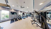 Located at 7560 Norman Rockwell Lane in Las Vegas, NV, this fitness center offers a comprehensive exercise environment designed to support a full range of workout routines. The space is thoughtfully laid out across a single floor, providing ample room for both cardio and strength training activities without feeling crowded.

The interior design features a distinctive geometric diamond-patterned accent wall in a navy and grey color palette, lending the space a modern, polished aesthetic. Light-toned flooring contrasts cleanly against the bold wall treatment, while a continuous decorative border near the ceiling ties the room together cohesively.

The cardio zone includes multiple treadmills and elliptical machines from TRUE Fitness, as well as stationary and recumbent bikes, all positioned to face large windows that invite natural light and outdoor views. Multiple flat-screen televisions are mounted throughout, allowing users to stay entertained during their workouts.

The strength training area is well-appointed with a multi-station cable machine system, free weights, and various resistance and functional training equipment. Full-length mirrors line one wall, enhancing the sense of openness and allowing users to monitor their form. Ceiling fans and recessed lighting ensure the environment remains comfortable and well-lit throughout. The overall ambiance is clean, modern, and purpose-built for fitness.