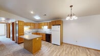 This single-floor home offers a practical and comfortable layout designed for everyday living. The kitchen is a standout feature, showcasing abundant warm-toned oak cabinetry that extends to the ceiling, offering generous storage throughout. A central island with a neutral countertop provides additional workspace, while light hardwood-style flooring adds a clean, bright aesthetic. White appliances, including a range and refrigerator, complement the space, and a decorative chandelier overhead adds a touch of character. The kitchen flows seamlessly into an open living and dining area, where plush carpeting, a ceiling fan with integrated lighting, and a second chandelier create a relaxed, welcoming atmosphere. The bedrooms are carpeted and well-sized, each benefiting from ceiling fans and natural light through double-pane windows with outdoor views. Closet storage is built in, maintaining a tidy and functional feel. The bathrooms feature warm wood vanity cabinetry, single-sink countertops, large wall mirrors with bar lighting, and durable stone-look tile flooring. One bathroom includes a full soaking tub, while another offers a half-bath configuration. Throughout the home, warm wood trim and neutral wall tones create a cohesive, understated design.
