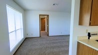 This single-floor residential space offers a clean and straightforward layout designed for comfortable everyday living. The interior features wall-to-wall carpeting in a neutral beige tone, providing a warm and quiet underfoot experience throughout the space. The walls are finished in a crisp white, creating a bright and open atmosphere that is easy to personalize. A textured ceiling adds a classic touch to the overall aesthetic. Natural light filters in through a window along one wall, contributing to the airy feel of the main room. A doorway with warm wood-toned trim leads into a connecting hallway, suggesting a thoughtful flow between the living areas. The hallway beyond provides access to additional rooms, hinting at a well-organized floor plan. Overhead, a ceiling vent ensures consistent climate control throughout. The space is accented by simple baseboards and standard electrical outlets, reflecting a no-frills, practical design. Overall, the space presents a neutral canvas well-suited for a variety of living arrangements, with its modest scale and clean finishes making it both functional and easy to maintain.