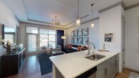 This thoughtfully designed single-floor residence offers a seamless blend of modern elegance and comfortable living. The open-concept layout connects the kitchen and living area, creating an inviting, airy atmosphere ideal for both everyday living and entertaining.

The kitchen is outfitted with gray shaker-style cabinetry, stainless steel appliances including a refrigerator, electric range, and built-in microwave, along with crisp white quartz countertops and a classic subway tile backsplash. A central island with seating provides additional prep space and a casual dining option, while pendant lighting adds a refined touch overhead.

The living area features tray ceilings with crown molding, warm hardwood-style flooring, and large windows with sliding glass door access that flood the space with natural light and offer views of the surrounding area. A gallery wall of marble-patterned art panels serves as a sophisticated focal point above the main seating area.

The bedroom is a tranquil retreat, highlighted by a bold slate-blue accent wall, plush carpeting, a ceiling fan, and generous natural light from a large window. The adjoining bathroom features a deep soaking tub framed by white subway tile walls and elegant navy drapery panels, evoking a spa-like sense of calm throughout.