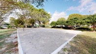 This outdoor recreational space centers around a well-maintained sand volleyball court, offering a dedicated area for sport and leisure. The court is bordered by concrete edging that defines its generous playing surface, with the sand providing an authentic and comfortable playing experience. A regulation net is installed across the center of the court, making it fully equipped for competitive or casual play.

Surrounding the court, mature trees with full canopies create a naturally shaded and visually appealing environment. The lush, green tree line encloses the space, offering a sense of seclusion while still maintaining an open, airy atmosphere. Benches are positioned nearby, providing seating for spectators or those looking to relax between games.

The open sky above allows for abundant natural light throughout the day, enhancing the welcoming and energetic ambiance of the space. The combination of natural greenery and recreational infrastructure makes this an inviting destination for both organized athletic activities and casual outdoor gatherings. Additional areas beyond the court suggest the presence of parking and further amenities, supporting the space's utility as a community or resort recreational facility.