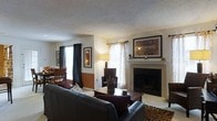 This single-floor residence offers a warm and functional layout centered around a well-appointed living area. The living room serves as the focal point of the space, anchored by a classic white-mantled fireplace with a dark surround, above which a large framed artwork adds a decorative touch. The seating area accommodates comfortable leather sofas and accent chairs arranged around a wooden coffee table, creating an inviting gathering space. A textured ceiling adds dimension overhead, while neutral-toned walls provide a versatile backdrop. Multiple windows dressed with layered curtains — combining sheer panels with dark drapes — allow for flexible light control while bringing in natural light throughout the day. A floor lamp and table lamp contribute to warm ambient lighting. Adjacent to the living area, a dining space is visible, featuring a table and chairs, suggesting a seamlessly connected open-plan arrangement. The flooring appears light-toned throughout, complemented by an area rug beneath the seating arrangement. Overall, the space presents a traditionally styled, comfortable interior well-suited for everyday living and entertaining.
