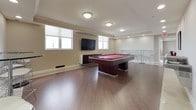 This well-appointed single-floor amenity space is designed for social gatherings and recreation, offering several distinct functional zones that flow cohesively together. The interior is finished throughout with neutral warm-white walls, crown molding, and a combination of large-format porcelain tile and warm wood-tone flooring, creating a clean and contemporary aesthetic.

The dining area features multiple round glass-top tables paired with sleek white upholstered chairs, illuminated by recessed lighting and flush-mount ceiling fixtures. One dining zone showcases a tray ceiling detail, adding architectural interest. Framed abstract artwork adorns the walls throughout, contributing a polished, curated feel.

A spacious lounge area provides modular white seating with bold red accent pillows arranged beneath a gallery wall of framed artwork, offering a relaxed setting for conversation. Adjacent to the lounge is a wet bar area featuring granite countertops, dark wood cabinetry, a stainless steel appliance, and clear acrylic barstools—ideal for entertaining.

The game room is a standout feature, centered around a full-size pool table with a red felt surface, flanked by large windows that invite natural light. French doors in the dining zone open to an outdoor area, further extending the space's entertaining potential.