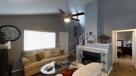 This two-story home in Arlington, Texas offers a thoughtfully designed living space that blends comfort and style. The main living area immediately impresses with its vaulted ceilings, which create an expansive, airy atmosphere and lend a sense of grandeur to the room. A ceiling fan with a light fixture is centrally mounted, providing both function and visual balance within the space.

The living room centers around a classic fireplace framed with a light-tiled surround, serving as a natural focal point and adding warmth to the room's ambiance. Decorative wall sconces and framed artwork add character to the soft gray-toned walls, contributing to an inviting, curated aesthetic.

Natural light flows in through a large window fitted with blinds, brightening the space while maintaining privacy. The room comfortably accommodates generous seating arrangements, ideal for both relaxing and entertaining.

A staircase with a white railing is visible in the background, leading to the second floor and reinforcing the home's multi-level layout. The overall design strikes a balance between casual comfort and polished detail, making this Arlington home a welcoming and functional residence.