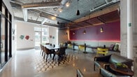 Located at 1601 Central Ave in St. Petersburg, Florida, this single-story space showcases a bold industrial-chic aesthetic that blends raw architectural elements with curated, colorful design details. Exposed concrete ceilings with visible ductwork and structural columns set the tone throughout, while polished concrete floors reflect abundant natural light streaming in through large glass entry doors and floor-to-ceiling windows that frame views of the surrounding greenery.

The main gathering area is a versatile, open layout anchored by a geometric-patterned area rug and a large communal dining table surrounded by mixed seating, including upholstered chairs in warm browns and grays. A built-in banquette along a deep burgundy accent wall—illuminated by pink neon underlighting—offers additional bar-height seating with yellow chairs, adding a vibrant pop of color. Pendant lights in matte black hang throughout, complementing the industrial framework above.

A separate, enclosed conference room features a substantial dark wood table, quilted gray chairs, glass-paneled walls, and black steel door frames, providing a quieter, more focused setting. The bright entry corridor serves as an informal gallery, displaying eclectic local artwork including sculptural wall pieces, paintings, and mixed-media works that reinforce the space's creative identity.
