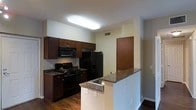 This single-floor residence offers a thoughtfully designed layout centered around a modern, fully equipped kitchen. The kitchen is a standout feature, showcasing rich dark wood cabinetry that provides ample storage, complemented by granite countertops with warm, earthy tones. Black appliances — including a gas range, over-the-range microwave, and a French door refrigerator — lend a sleek, cohesive aesthetic to the space. A peninsula with a built-in sink and matching granite surface extends into the living area, creating a natural divide between the kitchen and the adjoining space while also serving as a convenient breakfast bar or prep area. The hardwood flooring adds warmth and a sense of continuity throughout the unit. Overhead, a flush-mounted ceiling light fixture provides bright, even illumination across the kitchen. The neutral wall tones keep the space feeling open and versatile, allowing the darker cabinetry and countertops to take center stage. A interior door visible from the kitchen suggests a practical, connected floor plan. Overall, the space combines functional design with contemporary finishes for a comfortable and inviting everyday living experience.