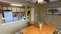 This single-floor residence offers a practical and comfortable layout centered around an open-concept dining and kitchen area. The dining space is anchored by a warm oak wood table with seating for multiple guests, accented by a decorative centerpiece. A ceiling fan with a light fixture hangs overhead, providing both airflow and ambient lighting to the room. Warm taupe-toned walls contribute to a cozy, inviting atmosphere throughout.

The kitchen is positioned adjacent to the dining area and is separated by a partial wall with a pass-through counter, maintaining a sense of openness while defining each functional zone. White cabinetry lines the kitchen walls, offering generous storage space, and is complemented by white appliances including a range and dishwasher. A built-in microwave and double sink with a window above add to the kitchen's functionality. Under-cabinet fluorescent lighting ensures a well-lit workspace.

A louvered bi-fold door, likely leading to a pantry or utility closet, is visible near the kitchen, adding practical storage. Dark hardwood-style flooring grounds the space and contrasts nicely with the lighter cabinetry and walls, creating a balanced, well-appointed interior.