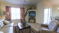 This single-floor space offers a thoughtfully designed living area that balances comfort and style. The living room serves as the centerpiece, anchored by a decorative corner fireplace with ornate metalwork detailing and accent pillows, above which a wall-mounted flat-screen television is neatly integrated into a built-in niche. A display shelf above the TV provides additional space for decorative accents, including a tall green bottle and dried botanical arrangements. The room features plush wall-to-wall carpeting that adds warmth underfoot, complemented by a patterned area rug beneath an elegant glass-topped side table with a wrought-iron base. Earthy tones dominate the decor, with cream-colored walls, warm brown drapery, and nature-inspired decorative pieces including large ceramic vases and tall dried grasses. Natural light fills the space generously through multiple windows fitted with horizontal blinds, including a sliding glass door that hints at an outdoor area beyond. A ceiling fan overhead ensures comfortable airflow throughout the room. The overall ambiance is warm and inviting, blending earthy, natural textures with tasteful furnishings to create a relaxed yet polished living environment.
