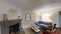 This single-floor space presents a thoughtfully designed interior that balances comfort and contemporary style. The living area serves as the heart of the home, anchored by a classic brick fireplace that adds warmth and character to the room. The textured ceiling adds architectural depth, while neutral-toned walls provide a versatile backdrop for the modern décor. The space is furnished with a plush gray sofa accented by blue and white throw pillows, complemented by a sleek glass-and-metal coffee table that reflects light and contributes to an open, airy feel. A patterned area rug in earthy tones grounds the seating arrangement, adding visual interest underfoot. Tasteful wall art, displayed as a pair of framed prints, enhances the curated aesthetic of the room. Tripod-style floor lamps flank the seating area, providing both ambient and accent lighting. Decorative wall sculptures and a small potted plant atop the fireplace mantel add finishing touches that give the space a lived-in yet polished character. The overall ambiance is warm, modern, and inviting, making it an ideal setting for relaxation and everyday living.