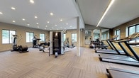This fitness center is thoughtfully designed to support a comprehensive workout experience within a clean, modern facility. The space is finished with neutral-toned walls and patterned carpet tile flooring throughout, creating a practical yet polished aesthetic.

The main workout area is generously proportioned, offering ample room to move between equipment without feeling crowded. Multiple treadmills are positioned along a windowed wall, providing natural light and outdoor views during cardio sessions. Elliptical trainers and stationary bikes complement the cardio offerings, while a multi-station strength machine anchors the center of the floor.

A dedicated free weights section lines one wall beneath large mirrors that visually expand the space. Dumbbells are neatly racked alongside a weight bench and stability ball, catering to a variety of strength and functional training routines. The mirrors also serve to maximize the sense of openness throughout the room.

Recessed ceiling lights and linear LED fixtures ensure consistent, even illumination across the entire facility. A secondary area near the entrance features wall-mounted displays, built-in shelving, and waste disposal stations, providing a convenient transition zone. Glass doors connect the space to an exterior access point, enhancing ventilation and accessibility. Overall, the fitness center is well-organized, bright, and fully equipped for diverse training needs.