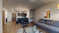 This single-floor unit is designed with a clean, modern aesthetic that balances functionality and comfort. The open-concept layout connects the living area and kitchen seamlessly, creating an efficient and cohesive living space. The living room features wood-style plank flooring, a sliding glass door that invites natural light and provides outdoor access, and enough room to accommodate a sofa, accent chair, and glass coffee table comfortably.

The kitchen is well-equipped with dark espresso cabinetry, white countertops, black appliances including a range and microwave, and a single-basin sink. A peninsula with bar seating serves as a natural divider between the kitchen and living area, adding both additional counter space and casual dining options. Pendant lighting above the peninsula enhances the kitchen's modern character.

The bedroom is carpeted for comfort and features a ceiling fan with light, a window allowing natural light, and ample space for a full bedroom set. The bathroom is finished with dark wood-tone cabinetry, a white countertop vanity, a large mirror, and wood-style flooring that ties the unit together with a cohesive design language throughout.