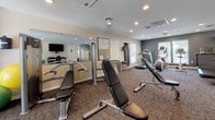 The Park Villas Apartment Homes community center at 21265 Mayfaire Lane in Lexington Park, Maryland, is a single-story facility thoughtfully designed to serve residents with both administrative and recreational amenities. The space is divided into two distinct functional areas: a leasing office and a fitness center.

The leasing office area features warm hardwood-style flooring throughout, recessed lighting, and neutral-toned walls that create a welcoming, professional atmosphere. A large reception desk with rich mahogany cabinetry serves as the focal point, complemented by upper storage units and space for administrative equipment. A resident computer station is also available. Natural light flows generously through multiple windows fitted with white horizontal blinds. A cozy seating nook with a round table and modern chairs is positioned near the windows, with a wall-mounted television above for added comfort.

The fitness center is carpeted and well-equipped, featuring treadmills, an elliptical, a stationary bike, a weight bench, and a rack of free weights. A wall of mirrors enhances the sense of space, while a mounted television and a dark accent wall add a dynamic, motivating aesthetic. Recessed lighting and additional windows ensure the area remains bright. An accessible restroom is conveniently located within the fitness area.
