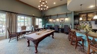 This well-appointed single-floor amenity space combines recreation, productivity, and comfort in a cohesive, elegantly designed environment. The main gathering area impresses with soaring ceilings, textured sage-green walls adorned with decorative medallions, and patterned carpeting that anchors the room. A full-sized billiards table takes center stage beneath a statement candle-style chandelier, while an adjacent games area features a dedicated chess and board game table surrounded by upholstered chairs. Large windows dressed with floor-length drapes flood the space with natural light and offer views of lush outdoor greenery, with French doors providing direct access to the exterior. A built-in business center lines one wall, offering multiple computer workstations illuminated by pendant lighting and framed artwork. The fully equipped kitchen showcases rich dark cabinetry, granite countertops, stone tile flooring, a stainless steel range topped by a striking copper hood, stainless steel dishwasher, and a tiled backsplash. A secondary prep area with a double sink, stainless steel dishwasher, and ample cabinetry completes the culinary offerings. Together, these areas create a versatile and inviting community hub well-suited for socializing, leisure, and work alike.