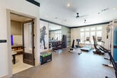 Located at 180 Hughes Rd in Madison, Alabama, this fitness center offers a thoughtfully designed exercise space across a single floor. The main workout area is bright and open, bathed in natural light from large multi-pane windows that frame views of the surrounding neighborhood. The room features durable rubber-backed flooring throughout, ceiling fans for air circulation, and recessed lighting to create a well-lit training environment.

The space is equipped with a comprehensive range of fitness equipment, including multiple treadmills, an elliptical trainer, a multi-function cable resistance machine, an adjustable weight bench, and a full dumbbell rack with weights displayed along a mirrored wall. Large floor-to-ceiling mirrors enhance the sense of space and allow users to monitor their form. A dedicated area accommodates a Echelon cycling bike with a mounted screen for interactive workouts. Built-in shelving along the accent-striped walls provides organized storage for accessories and mats.

A separate glass-windowed room serves as a private studio or gaming/media space, visible from the main floor. A water fountain station and stainless steel waste receptacles are conveniently positioned near the interior doorway. Distinctive wall murals featuring athletic silhouettes and abstract splatter designs add an energetic, motivating ambiance to the overall environment.