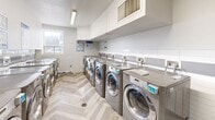 This shared laundry facility at 205–207 Morningside Ave in Toronto, Ontario, offers a clean, efficiently designed space for residential use. The room is lined on both sides with rows of modern stainless steel front-load washers and dryers, providing ample capacity for building residents. Each machine is equipped with a digital payment panel for convenient, cashless operation.

The interior features a bright, white color palette with crisp white cabinetry mounted overhead for storage, and a two-tone tile backsplash — white subway tile paired with grey tile — running along the walls to add a polished, contemporary touch. The herringbone-patterned light wood-look flooring adds a refined aesthetic to the utilitarian space.

Natural light filters in through a window along one wall, complementing the overhead fluorescent lighting to keep the room well-lit at all times. A stainless steel utility sink with a faucet is positioned along one wall, ideal for hand-washing or pre-treating garments. A cork bulletin board near the entrance displays posted notices and laundry instructions for residents. An electronic access panel is also present near the entry door. The room is neatly maintained, functional, and thoughtfully finished, making it a practical and comfortable shared amenity.
