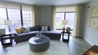 This single-floor living space exudes a contemporary, urban aesthetic defined by clean lines and a sophisticated neutral color palette of grey and gold. The living area is anchored by a curved charcoal sectional sofa adorned with patterned accent pillows, complementing the rounded grey ottoman coffee table that sits atop a plush white shag area rug. Dark hardwood flooring grounds the space and contrasts elegantly with the soft furnishings.

Natural light floods the room through large corner windows that frame views of a surrounding urban streetscape, creating a bright and airy atmosphere. The windows are dressed with bold geometric-patterned gold and white drape panels paired with neutral roller shades, offering both style and light control. A dark wood side table with a classic white lamp and framed artwork adds a warm, personal touch to the corner arrangement.

The walls are finished in a cool mid-tone grey, reinforcing the modern and cohesive design theme. A sculptural starburst decorative piece on the coffee table serves as an eye-catching focal point, adding dimension and visual interest to the overall composition. The space feels well-curated and inviting.