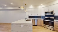 Located at 300 Enterprise Blvd in Bozeman, Montana, this well-appointed one-bedroom, one-bathroom residence offers a thoughtfully designed single-level layout with modern finishes throughout. The open-concept kitchen and living area serves as the heart of the home, anchored by a striking quartz waterfall island with an undermount sink and elegant gold pendant lighting. The kitchen features two-tone cabinetry — crisp white upper cabinets paired with warm wood lower cabinets — set against a dramatic dark stone backsplash. Stainless steel appliances, including a French door refrigerator, dishwasher, range, and microwave, complete the space. Light hardwood flooring flows through the main living area, which opens directly onto a private balcony through sliding glass doors, offering open views of the Bozeman skyline and surrounding neighborhood. The exterior showcases quality stone and siding detailing with wood-accented balcony ceilings. The bedroom is comfortably proportioned with plush carpeting, recessed lighting, and a natural window. A dedicated walk-in closet with wire shelving provides ample storage. Clean white walls and recessed lighting throughout create a bright, modern ambiance ideal for contemporary living.