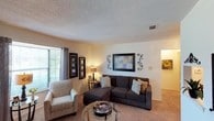 This single-floor residential space offers a warm and inviting atmosphere highlighted by a thoughtfully arranged living area. The living room serves as the heart of the home, featuring wall-to-wall plush carpeting that adds comfort underfoot and contributes to the cozy ambiance throughout. A large window dressed with elegant drapes allows generous natural light to fill the room, creating a bright and airy feel. The textured ceiling adds a classic architectural detail that complements the neutral wall tones. The space flows naturally from the living area into adjoining rooms, with open archways providing a seamless transition between areas. Decorative wall art and ornamental accents lend a personal and curated character to the interior. A glass-topped coffee table anchors the seating arrangement, which includes a plush sectional sofa and an accent chair, creating an ideal setting for relaxation or entertaining. Recessed ceiling vents suggest a well-integrated climate control system throughout. Overall, this space strikes a balance between functional design and comfortable living, making it a practical and welcoming environment well-suited for everyday life.