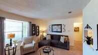 This single-floor residential space offers a warm and inviting atmosphere highlighted by a thoughtfully arranged living area. The living room serves as the heart of the home, featuring wall-to-wall plush carpeting that adds comfort underfoot and contributes to the cozy ambiance throughout. A large window dressed with elegant drapes allows generous natural light to fill the room, creating a bright and airy feel. The textured ceiling adds a classic architectural detail that complements the neutral wall tones. The space flows naturally from the living area into adjoining rooms, with open archways providing a seamless transition between areas. Decorative wall art and ornamental accents lend a personal and curated character to the interior. A glass-topped coffee table anchors the seating arrangement, which includes a plush sectional sofa and an accent chair, creating an ideal setting for relaxation or entertaining. Recessed ceiling vents suggest a well-integrated climate control system throughout. Overall, this space strikes a balance between functional design and comfortable living, making it a practical and welcoming environment well-suited for everyday life.