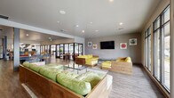 This single-story amenity center is thoughtfully designed to serve as a comprehensive community hub. The reception area sets an inviting tone with its warm wood-paneled ceiling, large-format light wood flooring, a substantial concrete-finished front desk, and floor-to-ceiling glass walls that draw in generous natural light and provide views of the surroundings.

The fitness center is expansive and well-appointed, divided into distinct zones. One area houses strength training machines, free weights, and benches alongside large windows overlooking the exterior. A separate section contains cardio equipment, including treadmills and ellipticals, positioned to take advantage of natural light. A dedicated functional training room features rowing machines, medicine balls, and stability equipment behind glass-panel partitions. Full-length mirrors enhance one zone, amplifying the sense of space.

The social lounge area features rich wood-tone flooring, bold green leather sectional sofas, and a full-size billiards table. Large windows frame outdoor views of a pool area. A yoga or stretching room with carpeted flooring and curated abstract artwork provides a quieter retreat. Throughout the facility, a consistent modern aesthetic is maintained with neutral tones, recessed lighting, and contemporary artwork.