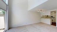 This two-floor space offers a thoughtfully designed layout that balances openness with functional living. The main living area immediately impresses with its dramatic double-height ceilings, creating a sense of expansive verticality that enhances the overall spaciousness of the room. Soft, neutral-toned carpeting runs underfoot, contributing to a warm and comfortable atmosphere. The open floor plan allows for flexible furniture arrangement, making the space highly adaptable to a variety of lifestyles.

Adjacent to the living area, the kitchen is both practical and clean in design. White cabinetry lines the walls from counter to ceiling, offering generous storage capacity. The countertops are finished in a light, neutral tone that complements the crisp white aesthetic throughout. A double stainless steel sink anchors the workspace, while a built-in dishwasher and additional appliances add everyday convenience. A track lighting fixture overhead ensures the kitchen is well-lit for cooking and entertaining. A pass-through counter connects the kitchen to the living area, promoting an open and social atmosphere. The second floor adds additional living space and further defines the multi-level character of this residence.